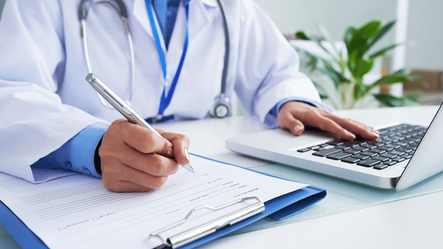 Close-up of a doctor in a white coat writing on a clipboard next to an open laptop computer.
