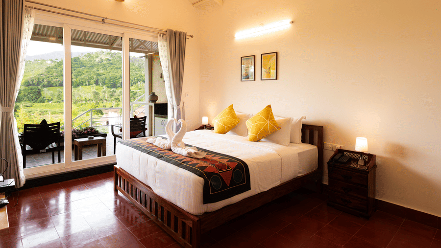 A bed with cushions and a runner sits on a polished floor inside the Bison room at Ibex Resort, Kotagiri. Glass doors with curtains lead to a balcony area. Frames hang on the wall above the bed structure.