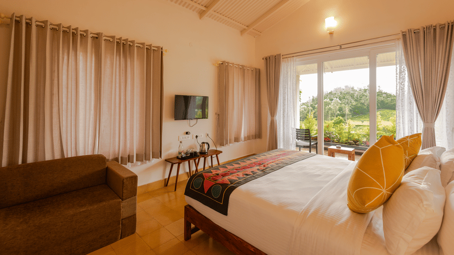 A bed with cushions and a runner stands inside the Bear room at Ibex Resort, Kotagiri. A television is mounted on the wall next to a curtained window. Glass doors at the back open towards an outdoor area.