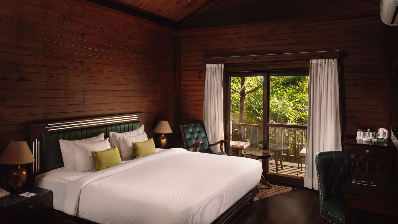 A corner view of Jungle Wood Bed Room  with a king sized bed, wooden walls, and bedside lamps and a spacious balcony.