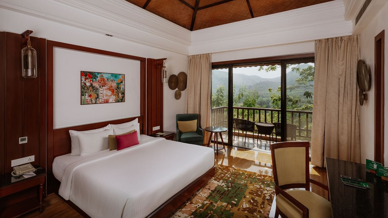 Corner view of the Luxury Villa bedroom at The Ananta, Udaipur, featuring a spacious balcony with scenic views, and a flat-screen TV, seating area with chair and a working desk.