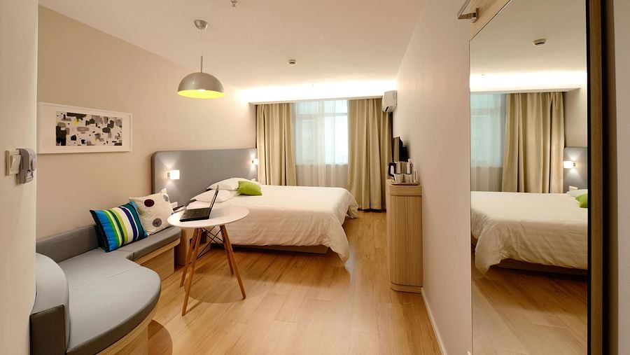 A bright, modern hotel room with a double bed, a curved sofa, and a small table with a laptop. A large mirror reflects part of the room.