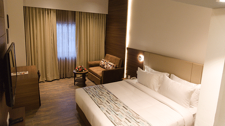A comfortable hotel room at Lords Inn Udaipur features a double bed with white linens, a cozy armchair by the window, and modern furnishings.