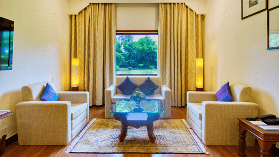 An Premium Suite living area at Clarks Khajuraho with armchairs, a center table, a television, and large curtains opening to a window view.
