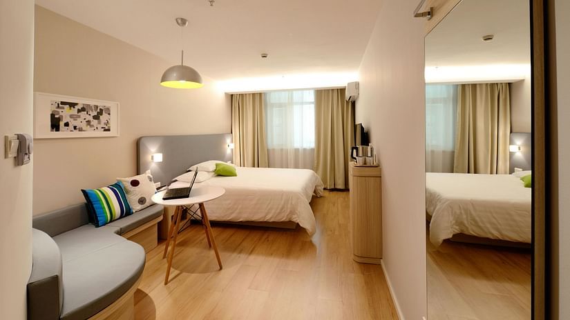 A bright, modern hotel room with a double bed, a curved sofa, and a small table with a laptop. A large mirror reflects part of the room.