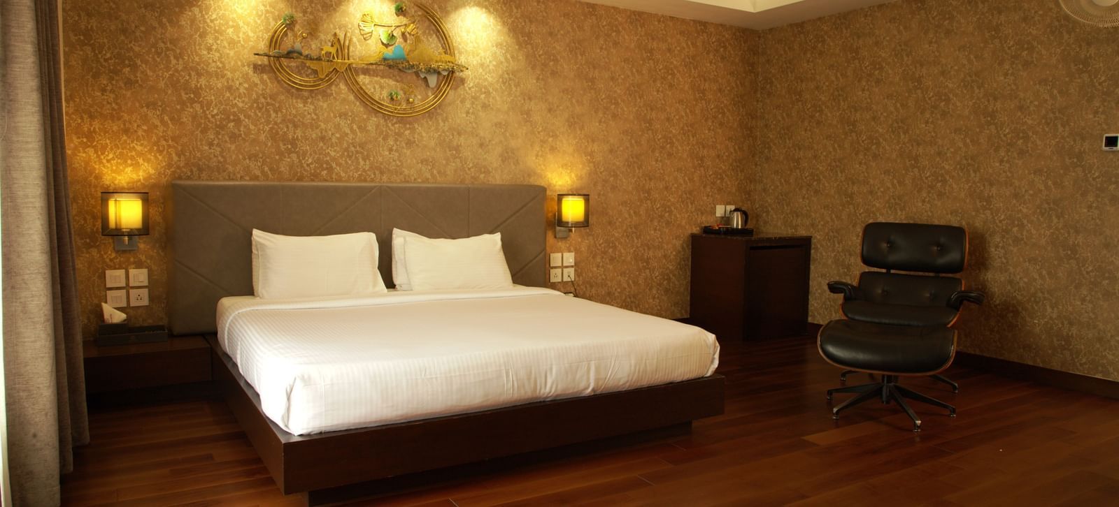 A warmly lit bedroom with a large bed, decorative wall panel, bedside lamps, and wooden flooring at The Hamuse Hotel, Kodaikanal.