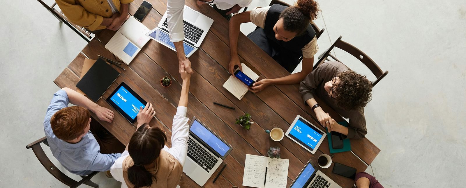 A high-angle shot where two individuals shake hands across a wooden table surrounded by other people, each with laptops or tablets