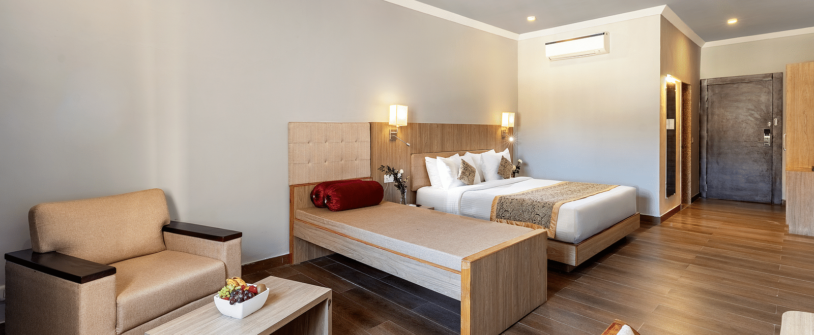 A well-lit hotel room with two beds, a beige armchair beside it and a small coffee table with fruits kept on it - Via Lakhela Resort & Spa, Kumbhalgarh