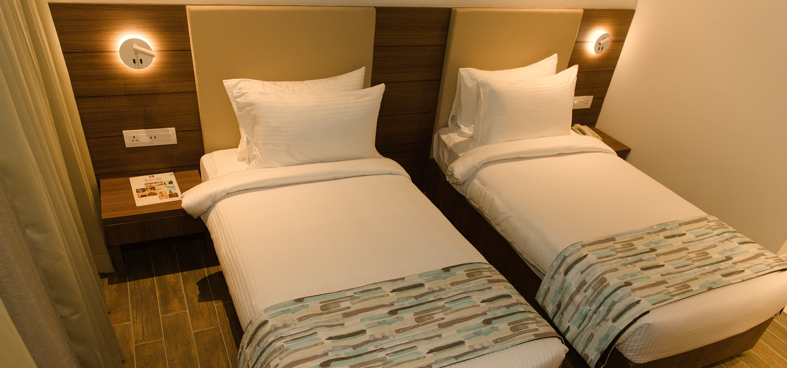A hotel room at Lords Inn Udaipur features two single beds with white linens and patterned runners, flanked by illuminated wall sconces.