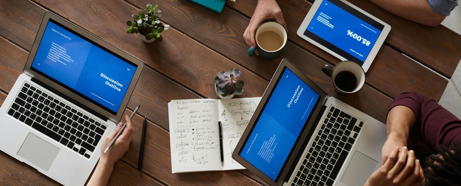A top shot of people sitting with open laptops placed on a brown table along with note books and cups of coffee.