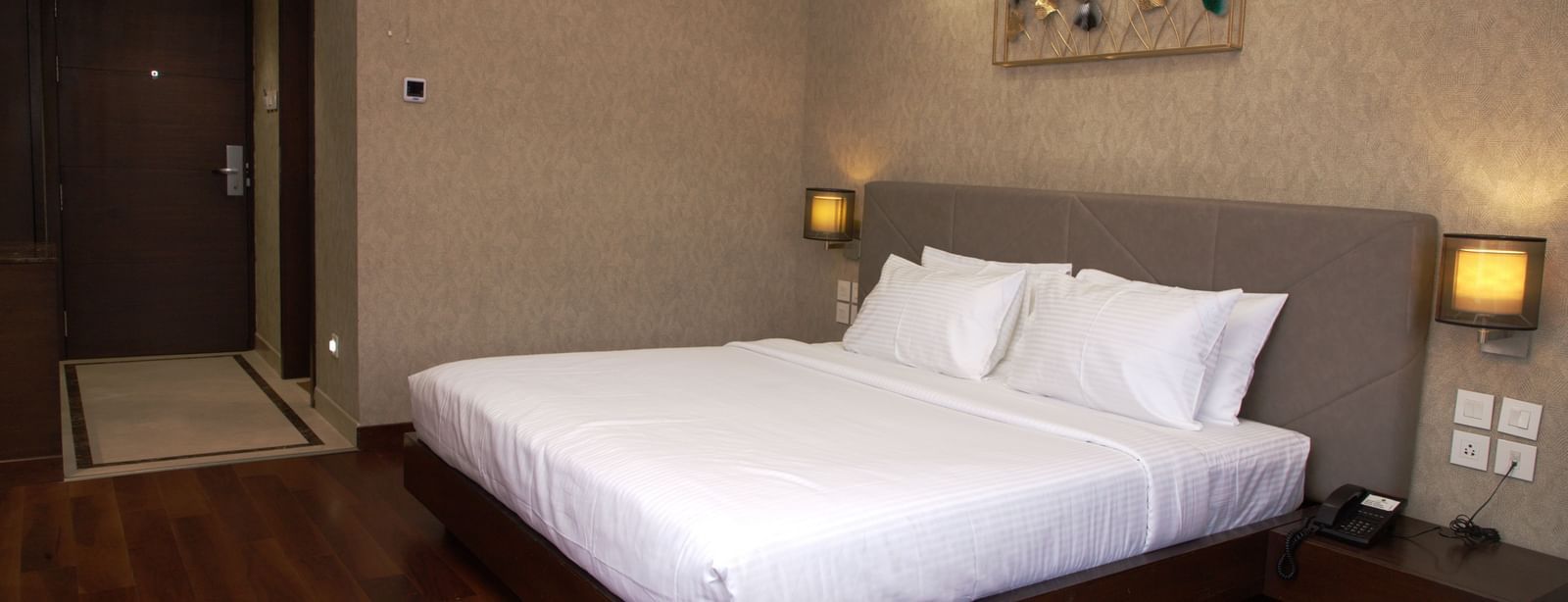 A spacious bedroom with a large bed, wall art above the headboard, bedside lamps, and wooden flooring at The Hamuse Hotel, Kodaikanal.