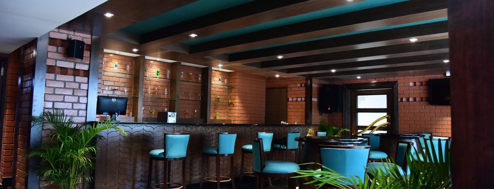 A vibrant hotel bar at The Royal Comfort, showcasing a brick-and-wood bar with bright blue-green barstools and a warm, inviting ambiance.
