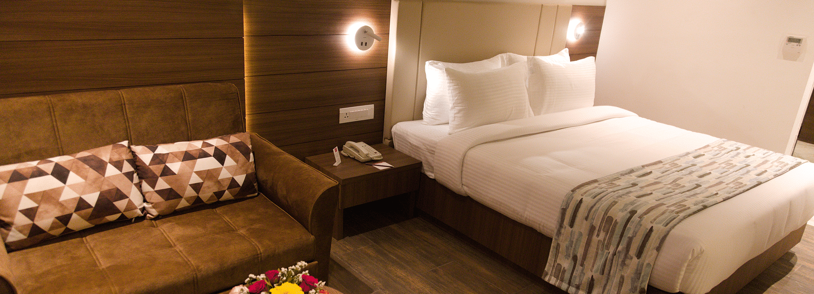 A well-appointed hotel room at Lords Inn Udaipur features a comfortable bed, a cozy sofa with patterned pillows, and a small round table with flowers.