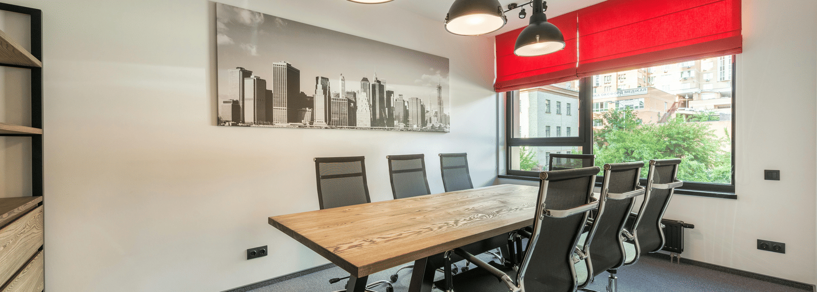 A modern conference room with a large wooden table, several black office chairs, hanging industrial-style pendant lights, and a window with red blinds.