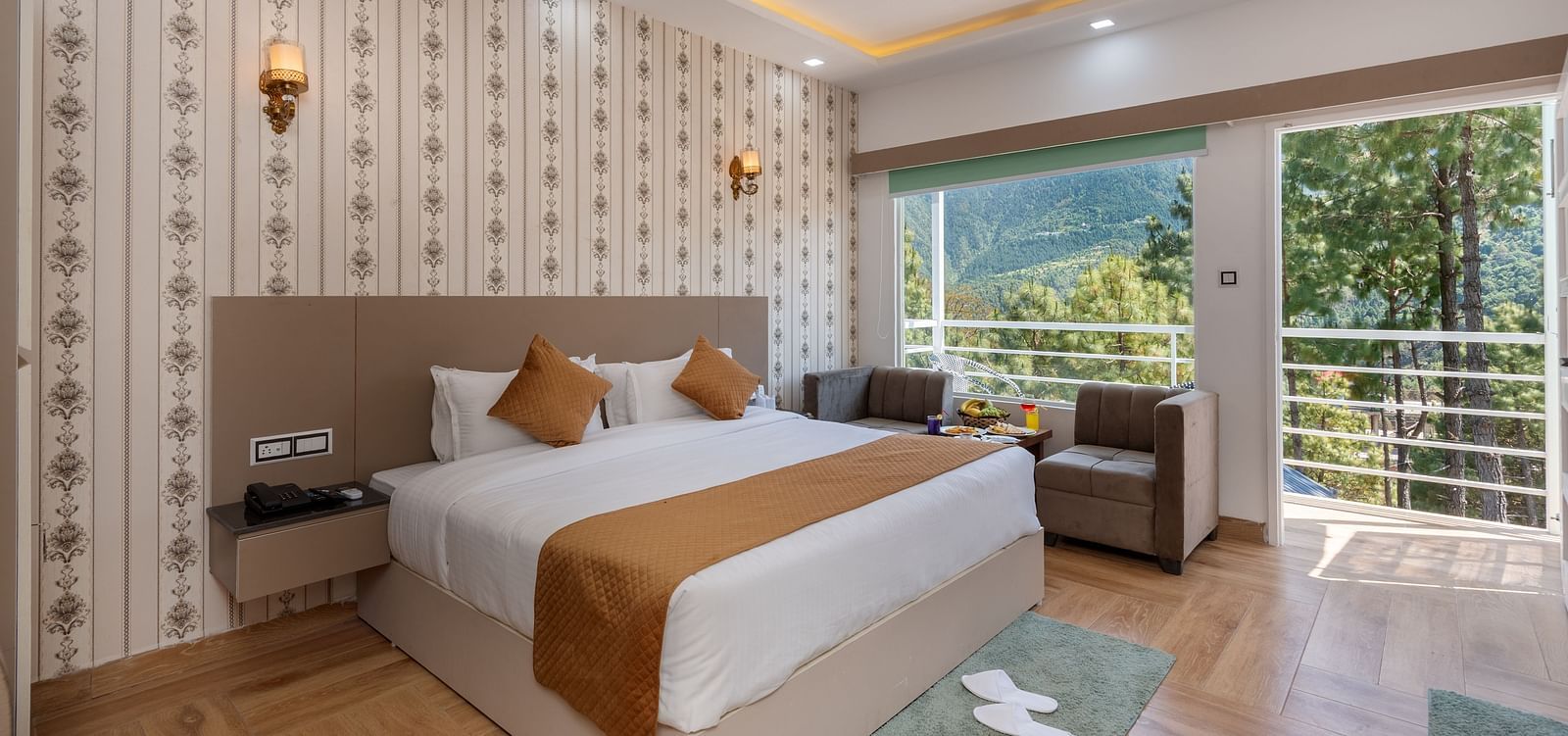 Spacious hotel bedroom featuring a large double bed with crisp white linens and brown accents, stylish wallpaper, a ceiling fan, modern lighting, a cosy seating area, and a balcony offering scenic views of lush greenery and hills - DLS Nature Trinket Resor