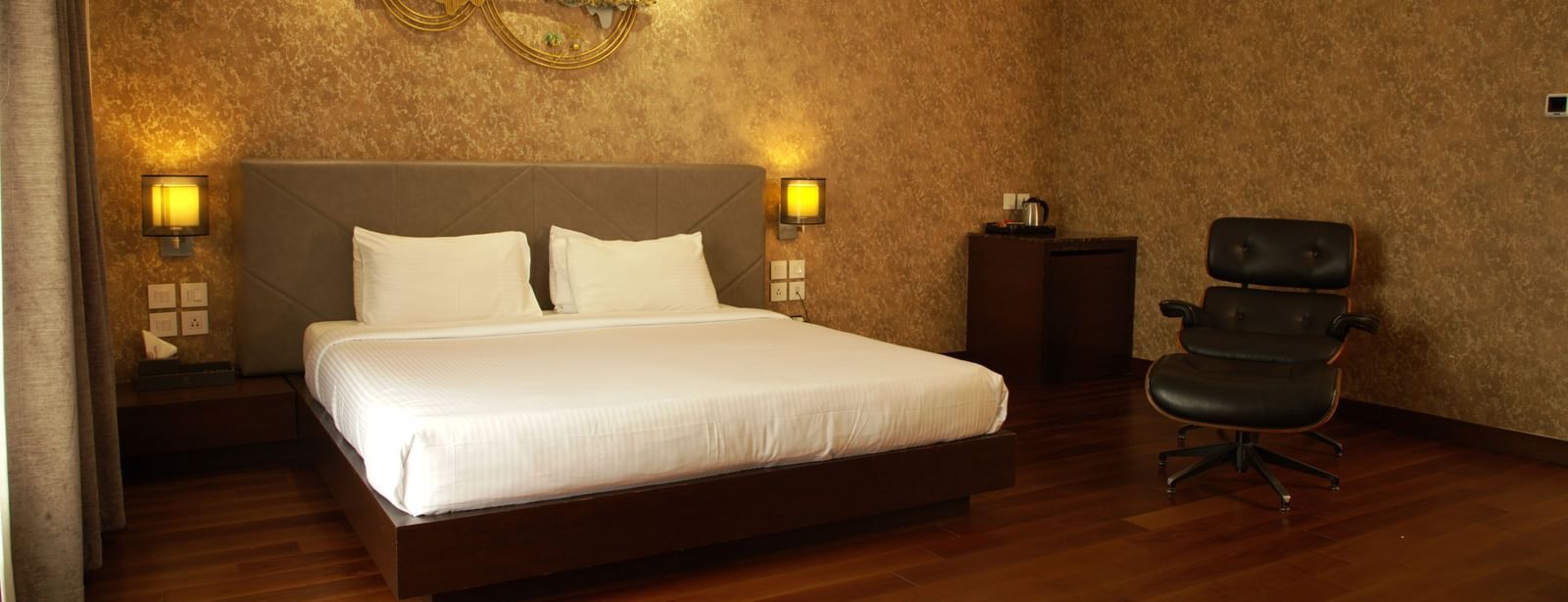 A warmly lit bedroom with a large bed, decorative wall panel, bedside lamps, and wooden flooring at The Hamuse Hotel, Kodaikanal.