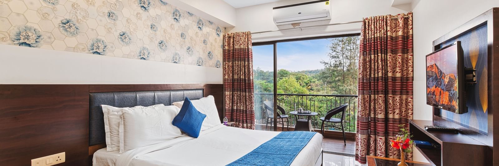 Bright and ventilated room at Mayur Retreat with a blue-themed bed, floral walls, and balcony garden view.