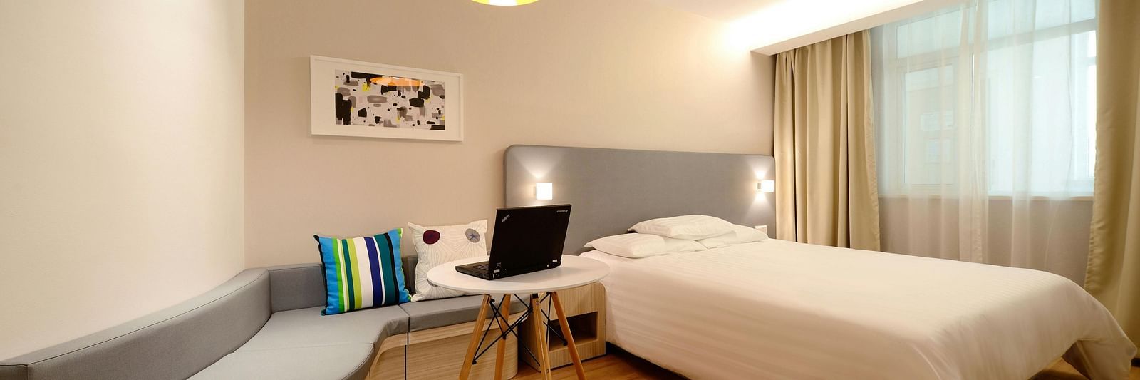Comfortable hotel room featuring a bed, built-in curved sofa, small round table with a laptop, and pendant lighting.