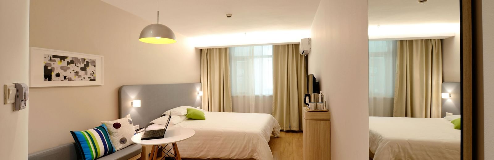 Modern hotel room with a bed, a curved sofa with colourful pillows, a small table, and a mirrored wall reflecting the bed.