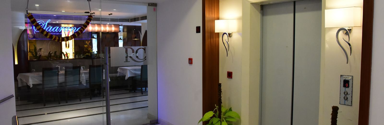 A sleek, modern elevator lobby at The Royal Comfort, with a metallic elevator door and glowing blue lights beneath a wooden ceiling panel.