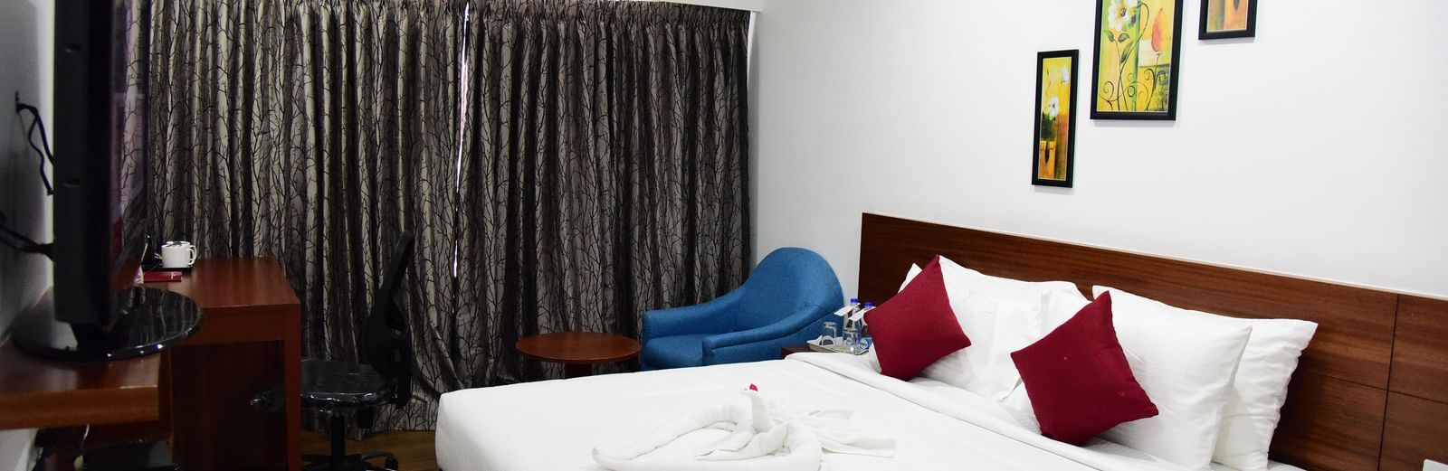  A spacious royal deluxe room at The Royal Comfort, featuring a king-sized bed with rich wooden accents, a deep red runner, and a modern painting above the headboard.