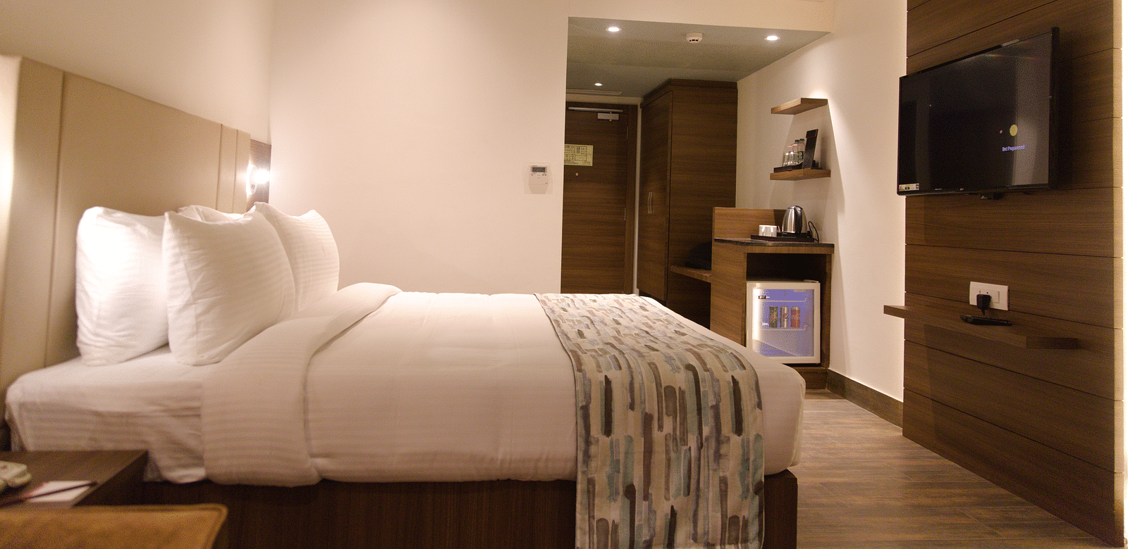 A comfortable hotel room at Lords Inn Udaipur is furnished with a double bed, a mounted television, and a small refrigerator unit.