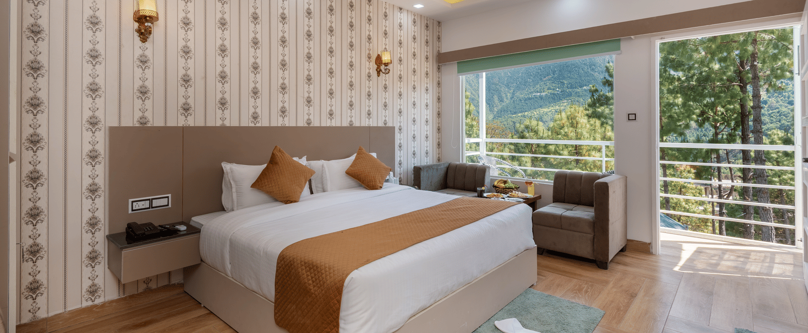 A spacious hotel bedroom featuring a large double bed with crisp white linens and brown accents, a cosy seating area, and a balcony offering scenic views of lush greenery and hills - DLS Nature Trinket Resort, Dalhousie