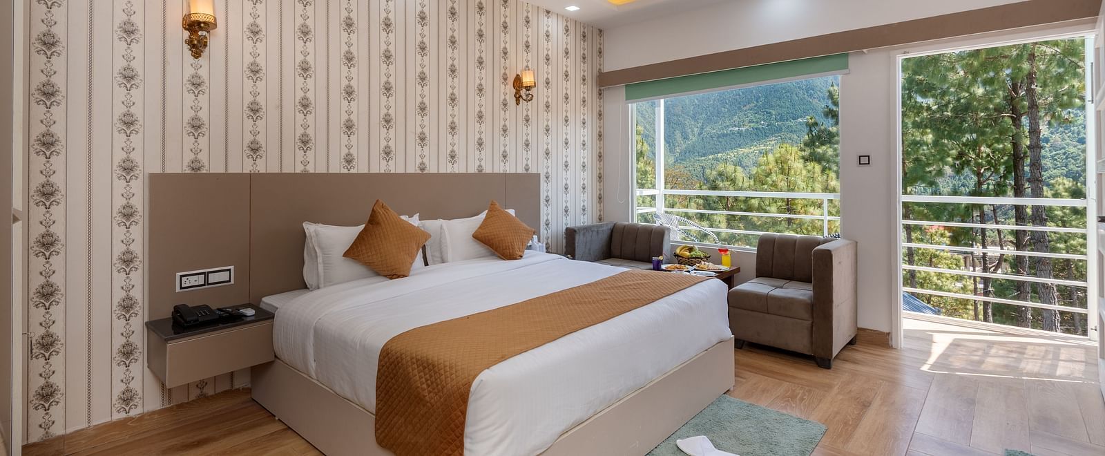 Spacious hotel bedroom featuring a large double bed with crisp white linens and brown accents, stylish wallpaper, a ceiling fan, modern lighting, a cosy seating area, and a balcony offering scenic views of lush greenery and hills - DLS Nature Trinket Resor