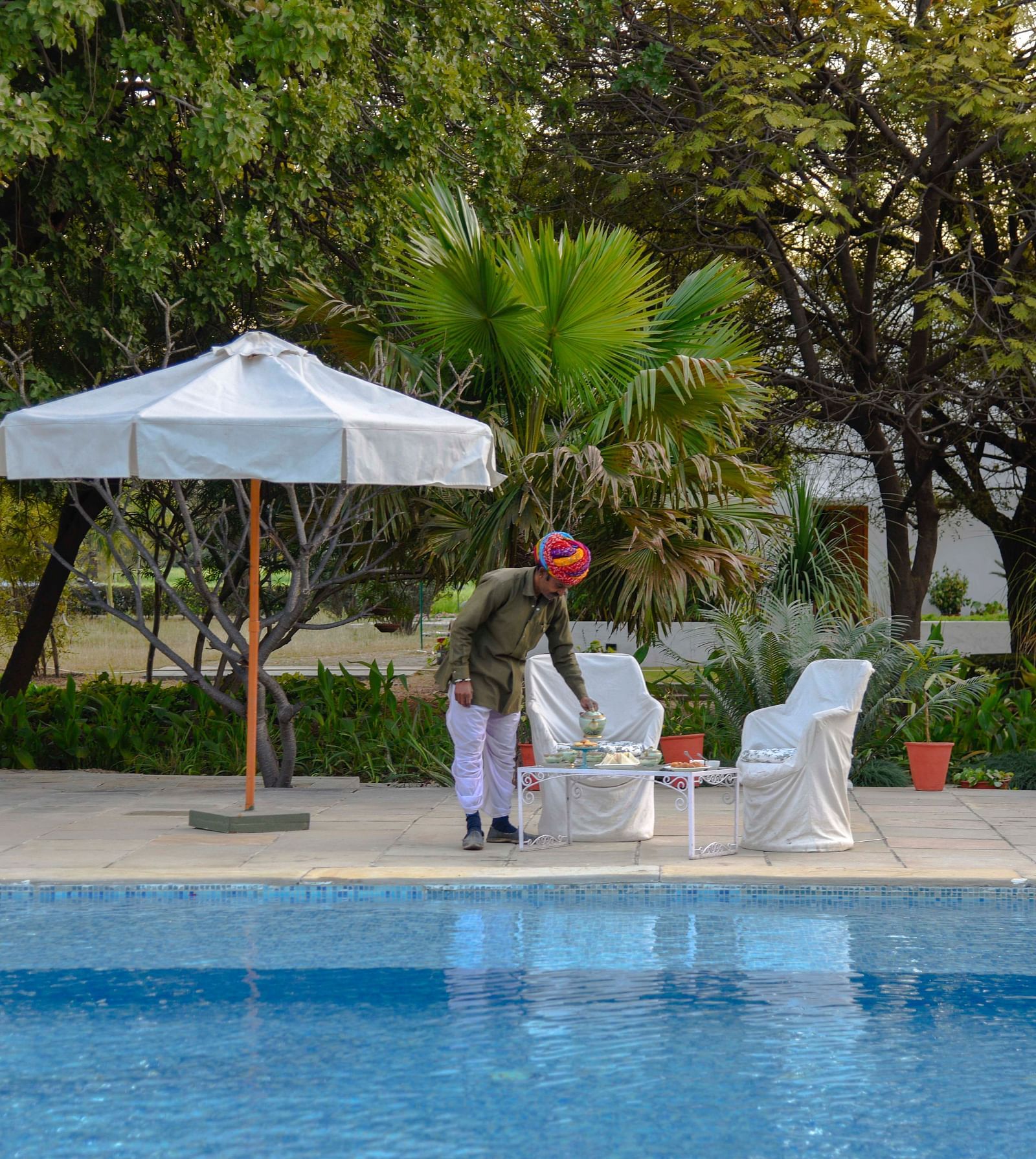 A person in a turban arranges items on a table beside a swimming pool, with a parasol and seating nearby, amidst lush greenery- Shahpura Bagh.