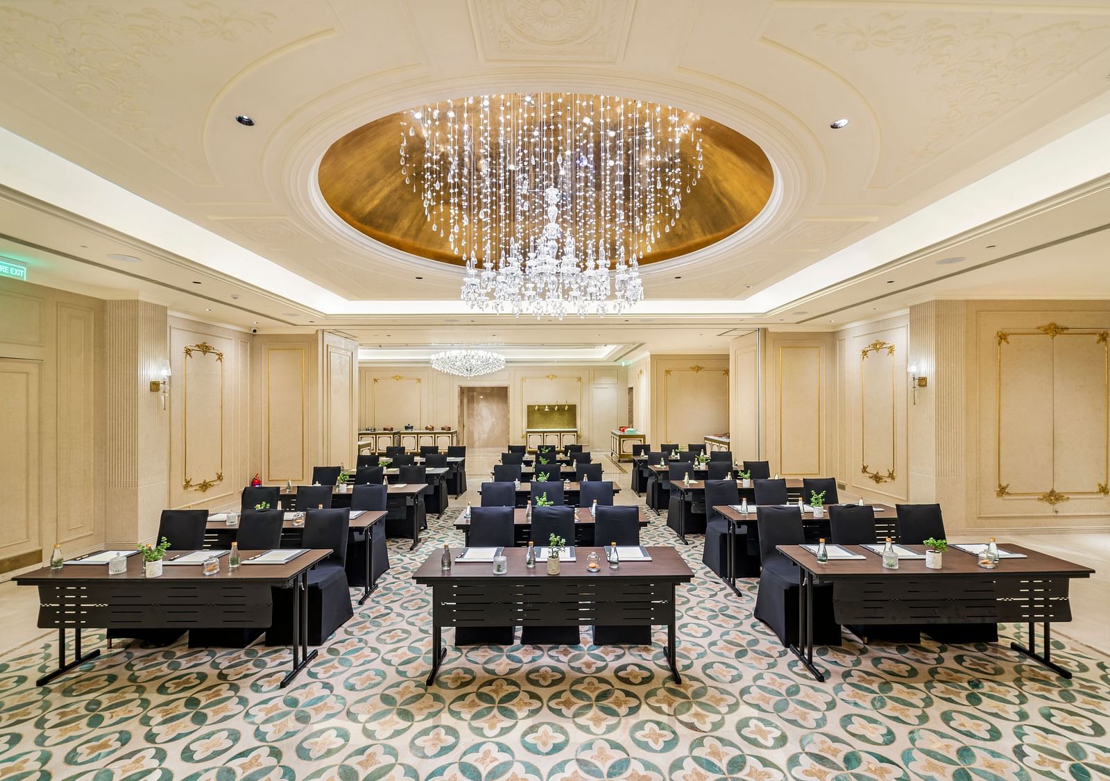 A grand ballroom configured for a large seminar or presentation at Grand Chennai by GRT Hotels showcasing numerous black tables and chairs neatly arranged in rows. A magnificent, large chandelier dominates the high ceiling, enhancing the luxurious feel.