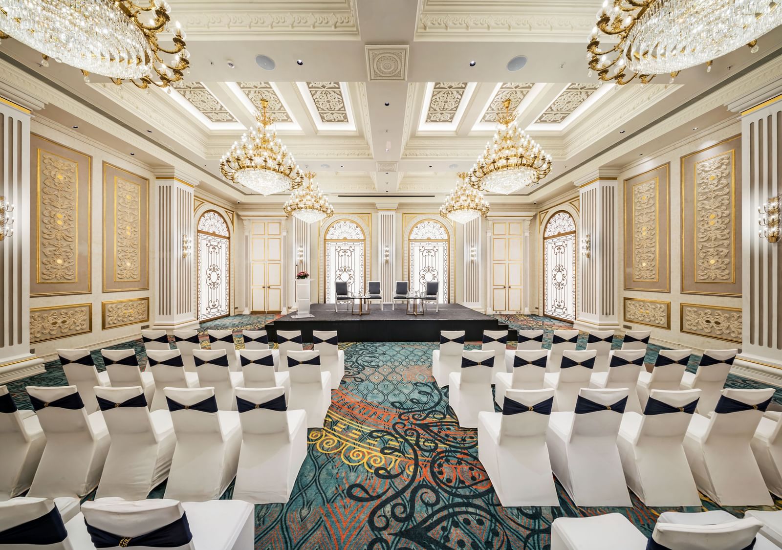 A beautifully adorned event hall set up for a presentation or ceremony at Grand Chennai featuring rows of white upholstered chairs facing a central stage. The intricate wall paneling and ornate ceiling fixtures contribute to a lavish setting.