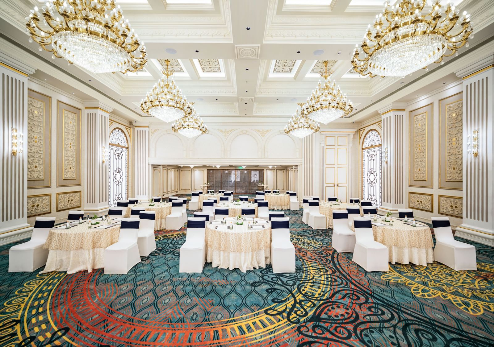 A spacious ballroom arranged with various white-draped tables and chairs at Grand Chennai ready to accommodate a diverse event setup, bathed in the soft glow of exquisite chandeliers.