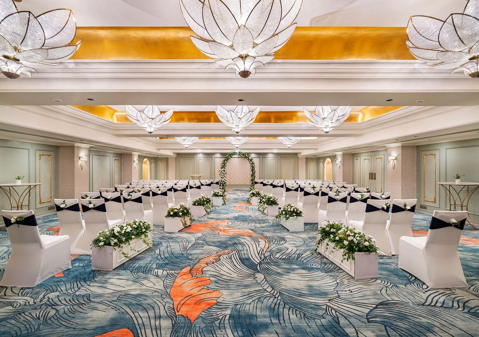 An elegant ballroom at Grand Chennai by GRT Hotels is set for a formal gathering, showcasing meticulously aligned white chairs, tasteful floral centerpieces, and magnificent, decorative chandeliers that illuminate the entire space.