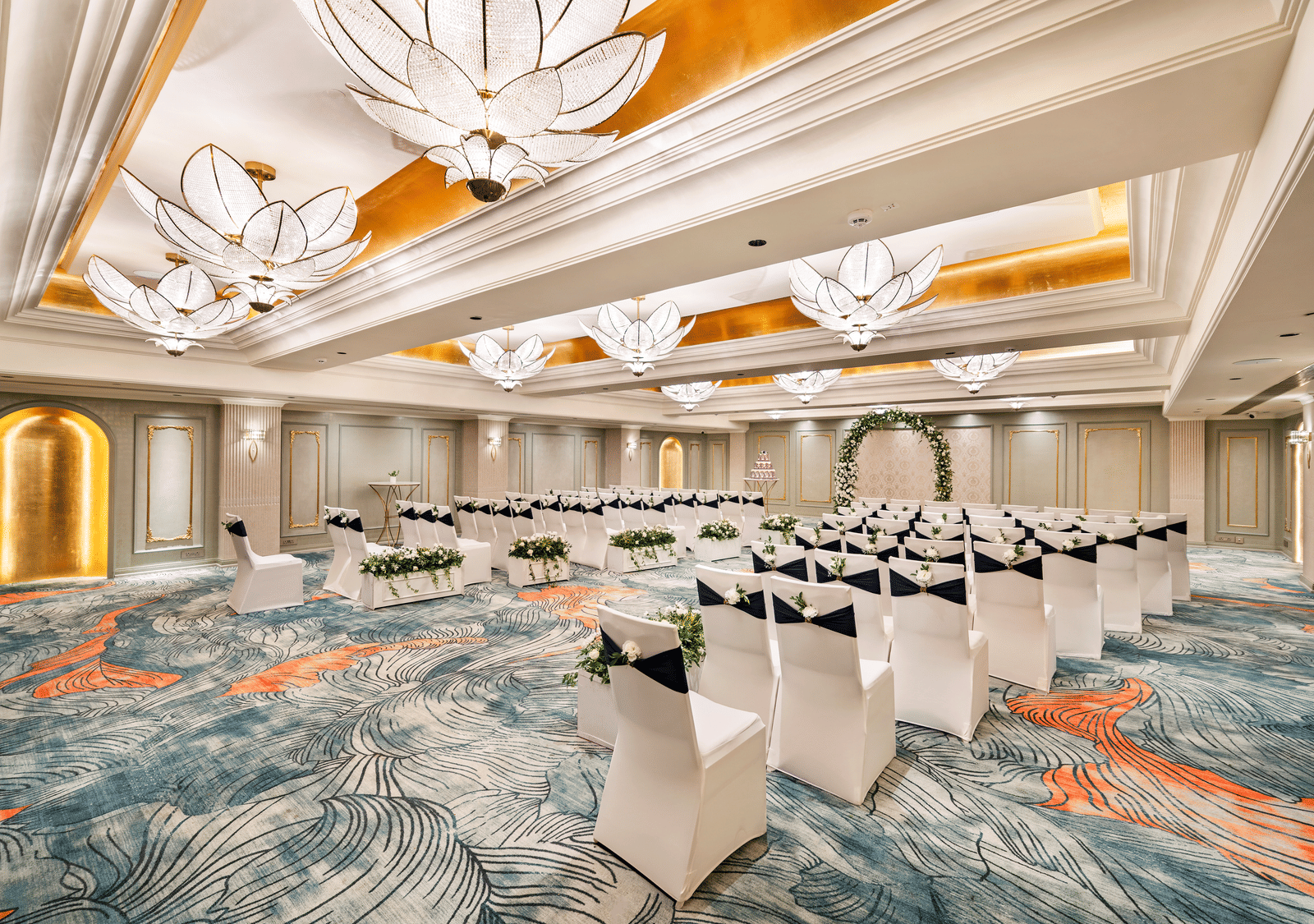 A grand and versatile event space at Grand Chennai by GRT Hotels, meticulously arranged with numerous white chairs facing a distant stage. The intricate patterned carpet and stunning ceiling fixtures enhance the room's luxurious ambience.