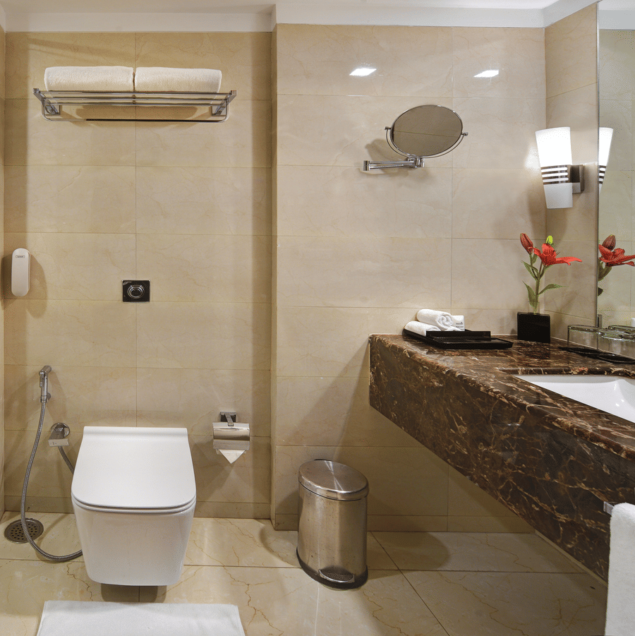 Bathroom at The Bristol Hotel, Gurgaon with beige marble tiles, a glass-enclosed shower, a wall-mounted toilet, and a dark marble vanity counter with a mirror.