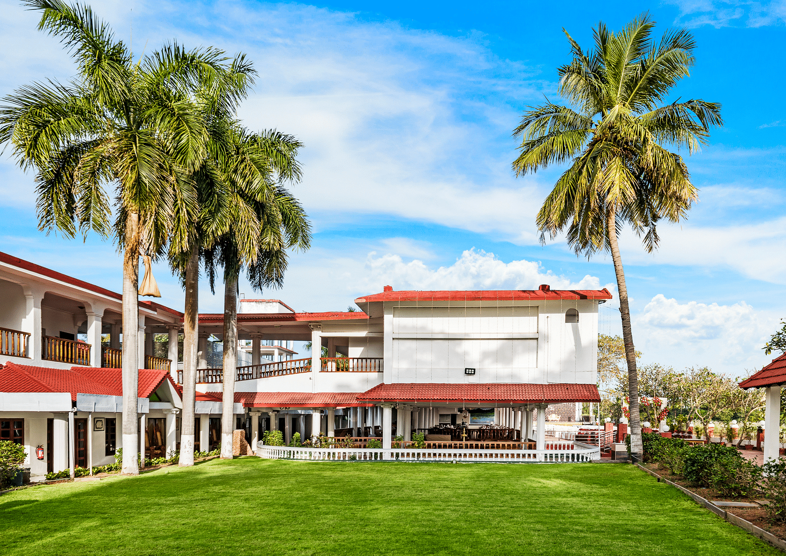 A facade image of the exterior view showcasing a green lawn, tall palm trees with the hotel infrastructure in the background - GReaT Trails River View Resort Thanjavur By GRT Hotels