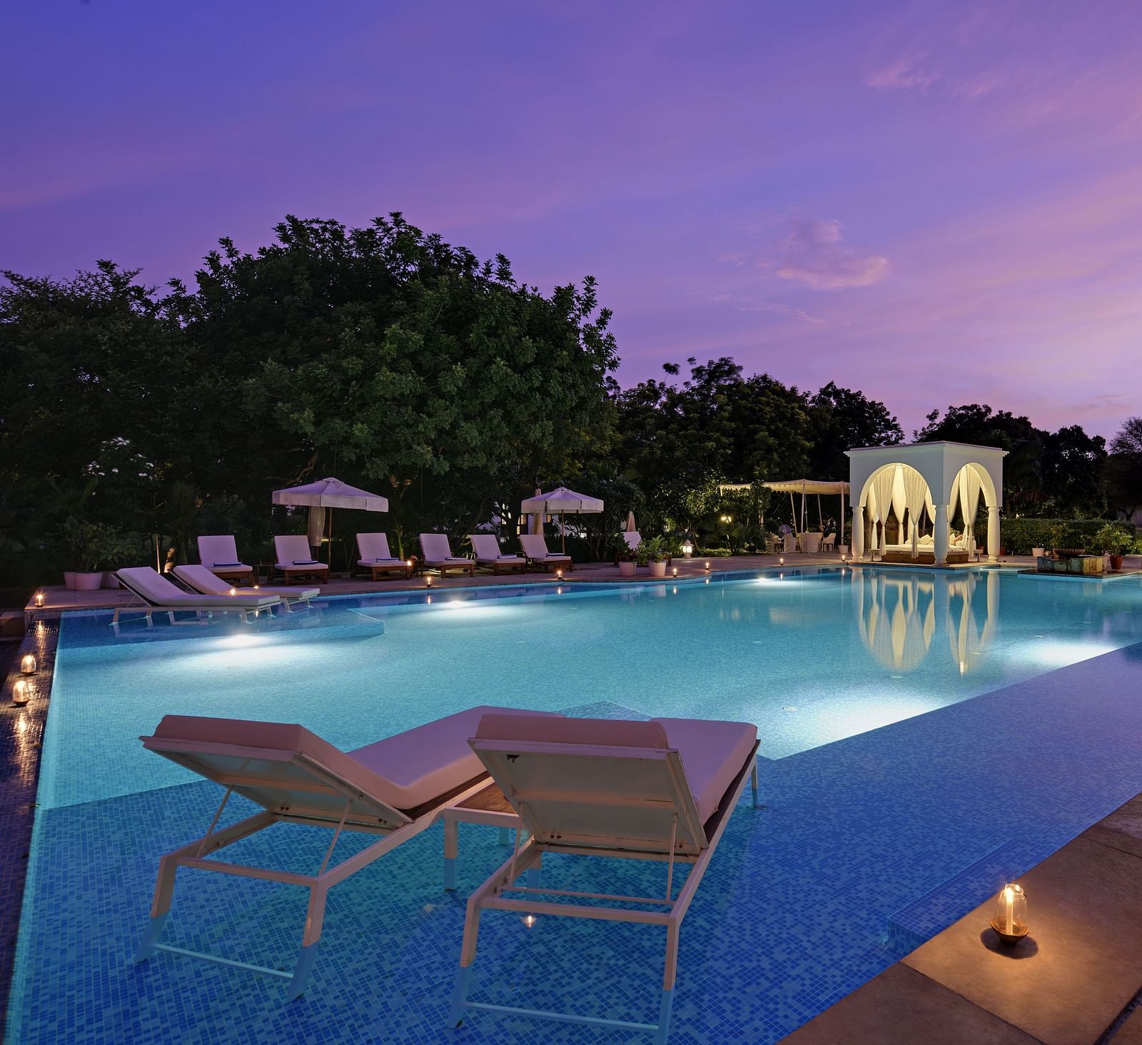 A serene swimming pool at dusk, flanked by lounge chairs and white structures with draped fabric, under a twilight sky- Shahpura Bagh.