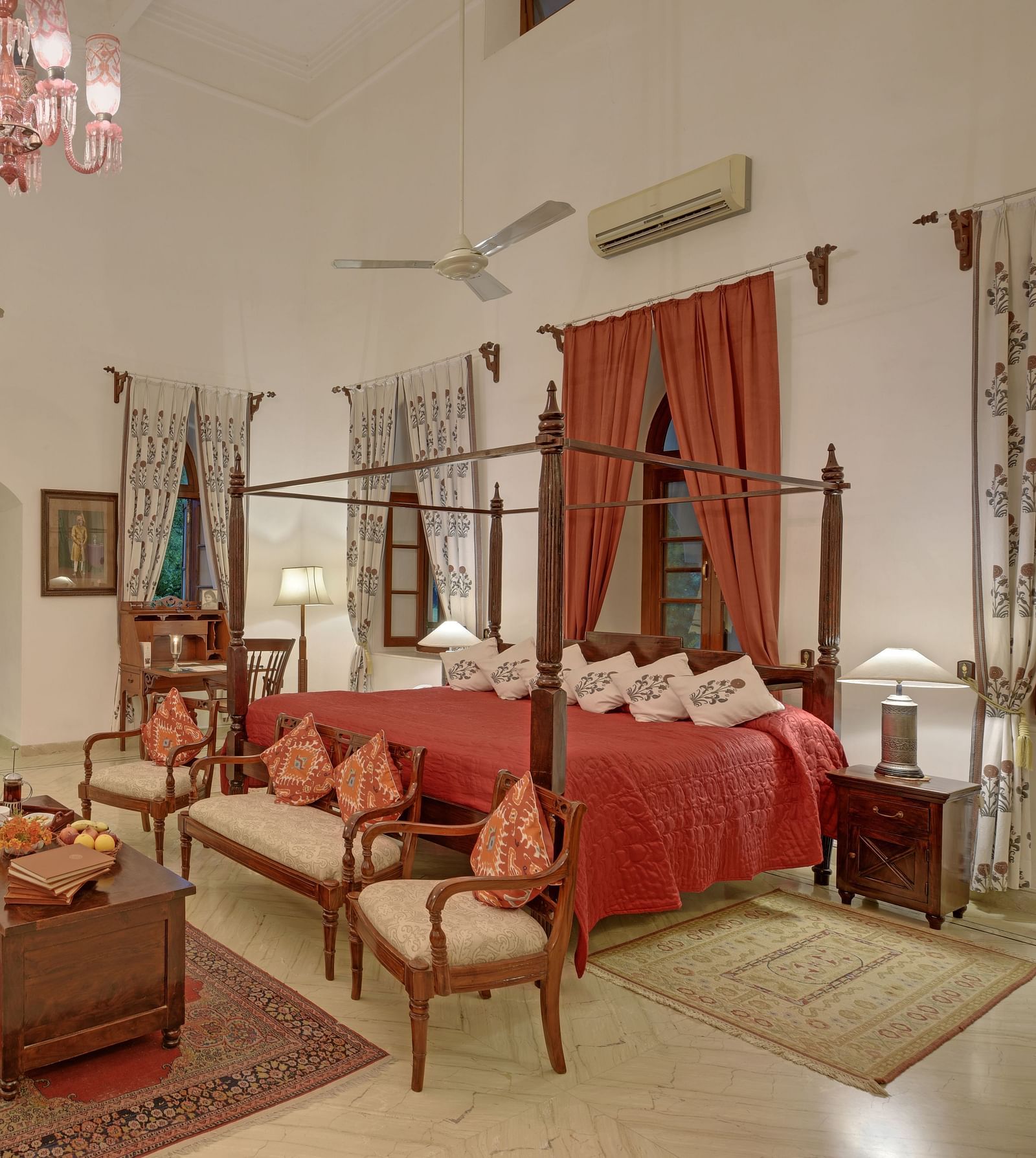 A well-appointed room with a four-poster bed, seating area, and decorative rugs, featuring arched windows and a chandelier, under a high ceiling- Shahpura Bagh.