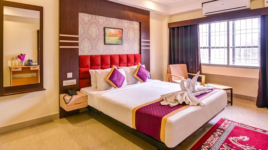 A bright hotel room with a red cushioned headboard, white bedding with purple and gold accents, a large window with curtains, and a mirror beside a wooden dressing table - Mount Hotels