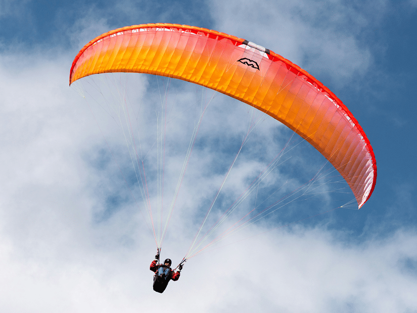 Paraglider flying beneath a bright orange canopy against blue skies, with soft white clouds and rugged mountain terrain far below