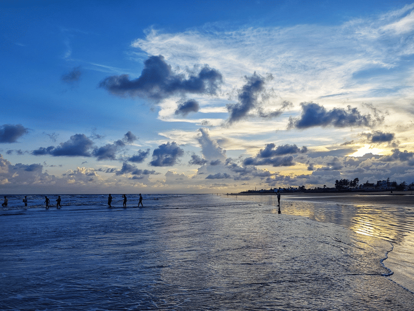 Visitors walk along the vast, wet sandy shores of Mandarmani Beach while the sun reflects brightly across the calm sea waves.