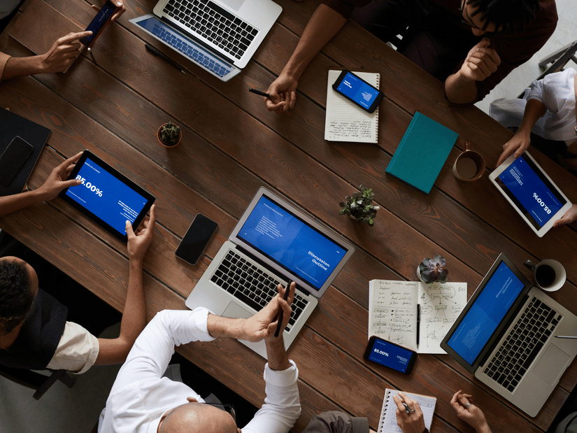 A high-angle view of several people gathered around a conference table working on laptops and phones during a meeting.