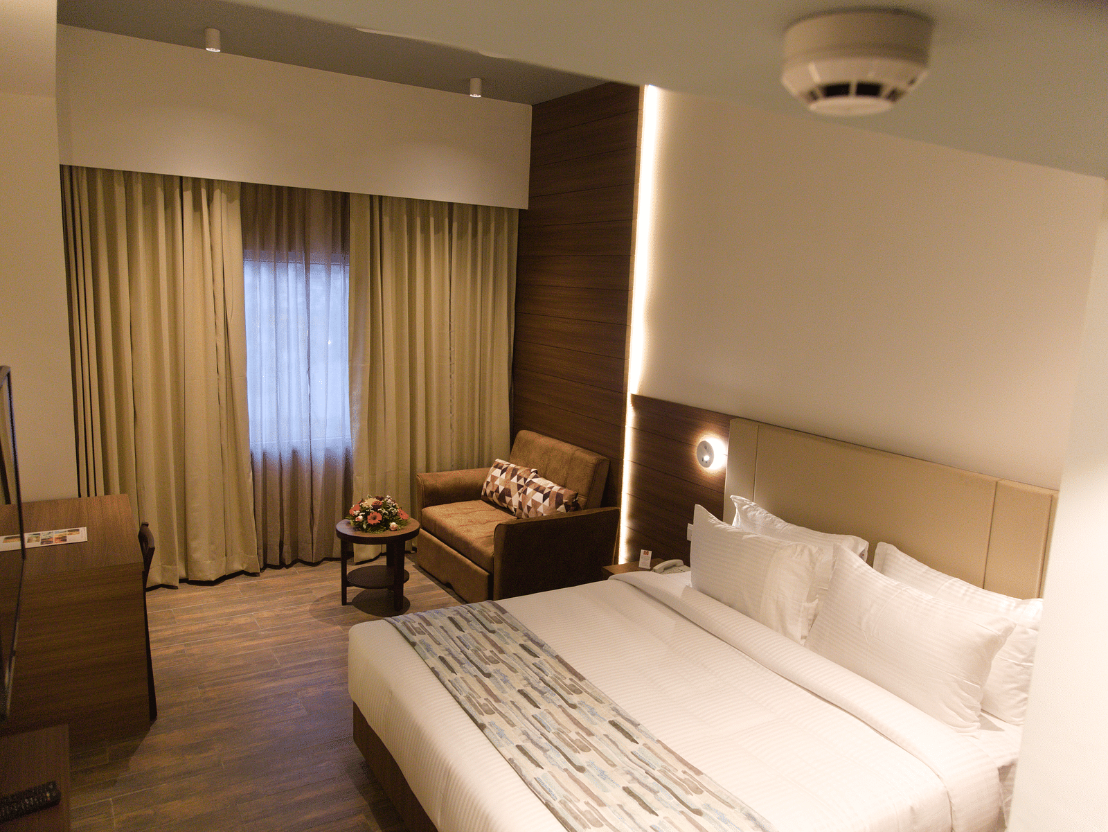 A comfortable hotel room at Lords Inn Udaipur features a double bed with white linens, a cozy armchair by the window, and modern furnishings.