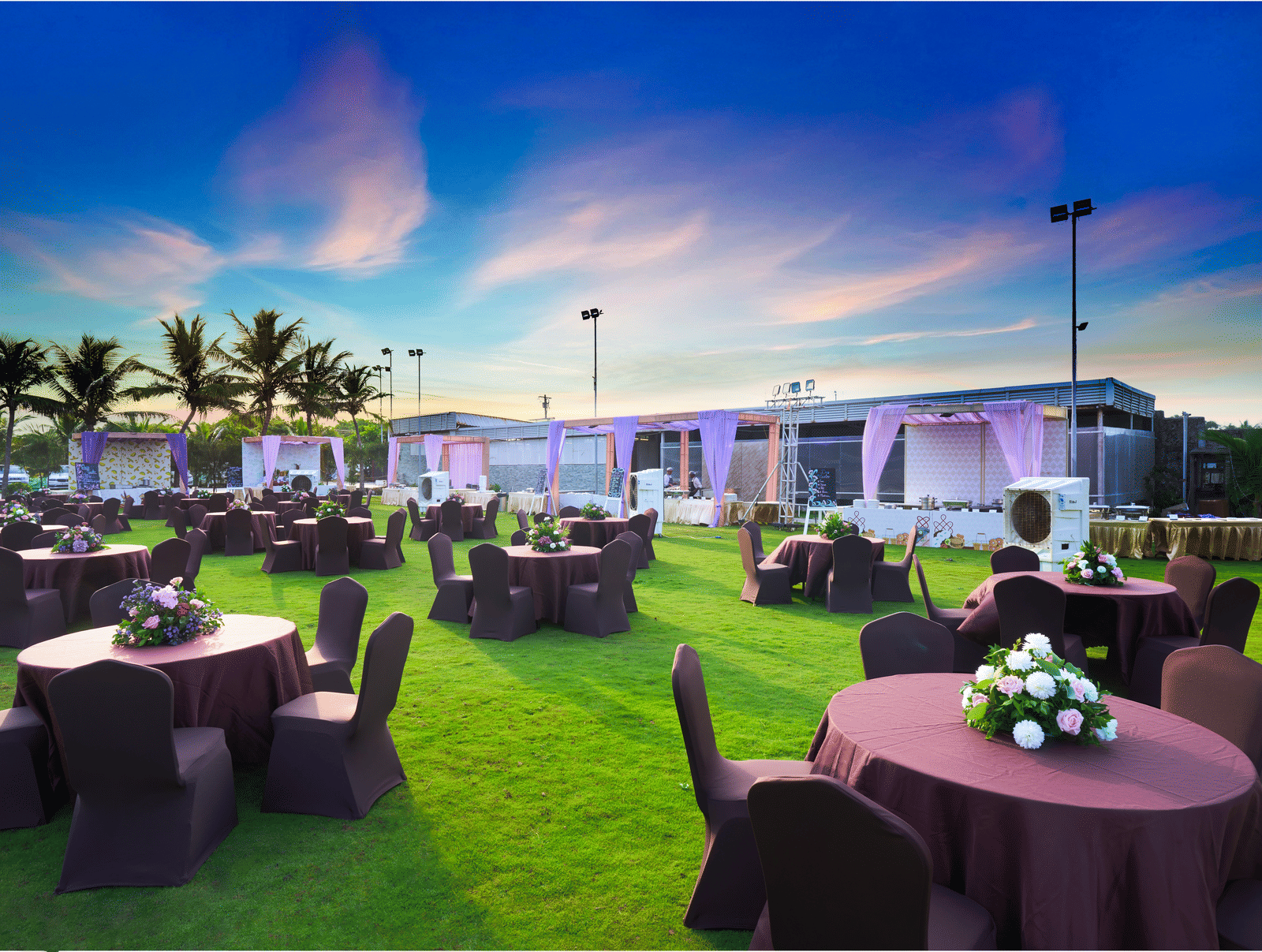 Outdoor lawn setup with round tables and floral centrepieces under a vibrant evening sky - Grande Bay Resort & Spa, Mamallapuram