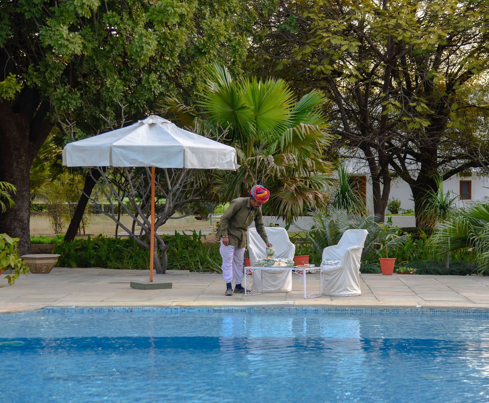 A person in a turban arranges items on a table beside a swimming pool, with a parasol and seating nearby, amidst lush greenery- Shahpura Bagh.