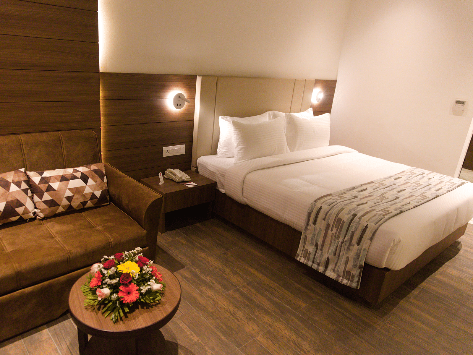 A well-appointed hotel room at Lords Inn Udaipur features a comfortable bed, a cozy sofa with patterned pillows, and a small round table with flowers.