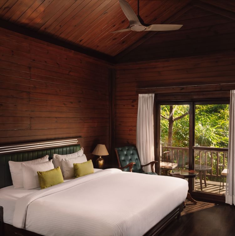 A corner view of Jungle Wood Bed Room  with a king sized bed, wooden walls, and bedside lamps and a spacious balcony.
