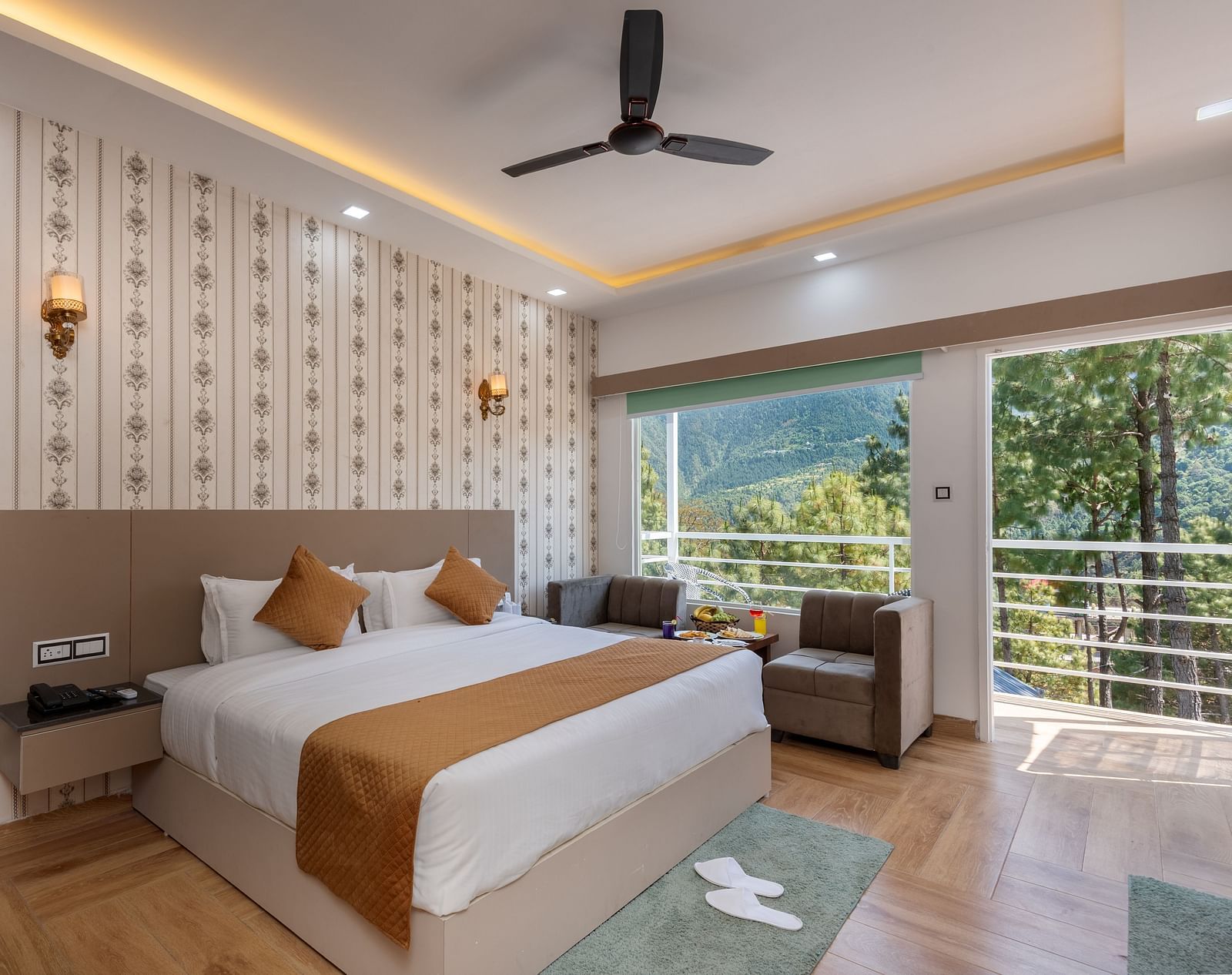 Spacious hotel bedroom featuring a large double bed with crisp white linens and brown accents, stylish wallpaper, a ceiling fan, modern lighting, a cosy seating area, and a balcony offering scenic views of lush greenery and hills - DLS Nature Trinket Resor