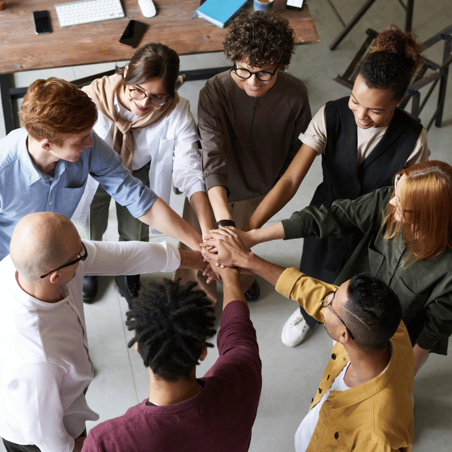 Group of eight people standing in a circle with hands joined together in the center, symbolising unity, teamwork, and collaboration in a professional indoor workspace.