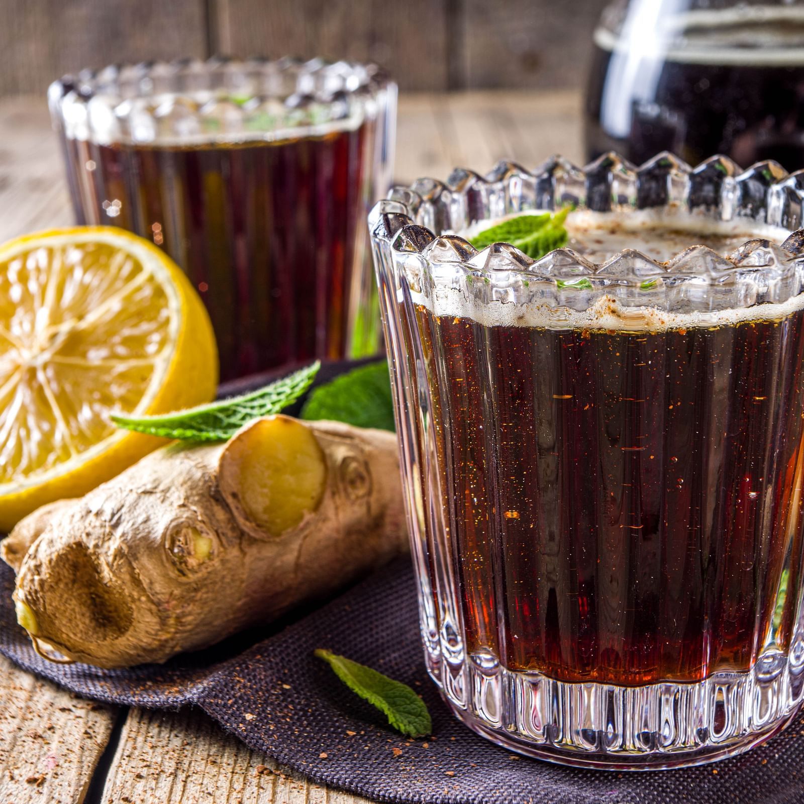 Two glasses of dark, frothy ginger beer with fresh ginger and orange slices on a rustic wooden table.